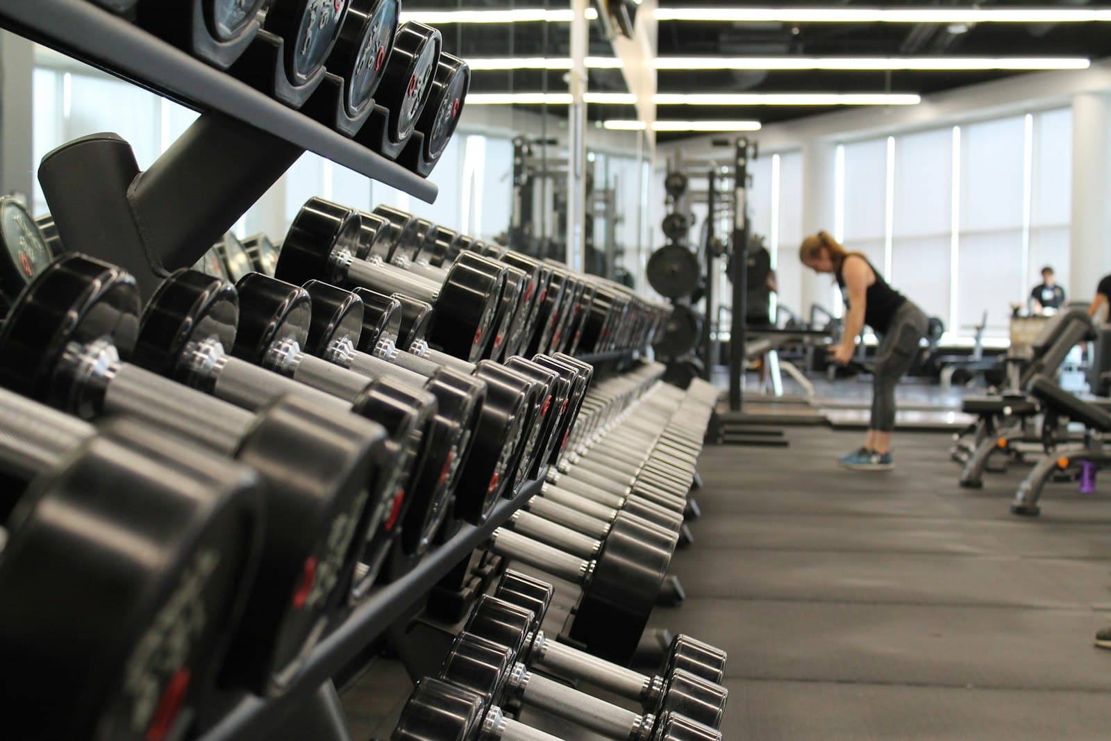 Rows of dumbbells on a rack in a bright gym, a woman training in the background